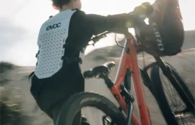 Persona con equipo de protección, incluido un EVOC Torso Protector talla XS para seguridad, lleva una bicicleta de montaña naranja cuesta arriba por terreno arenoso bajo la luz del sol.
