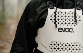 Persona vestida con una sudadera con capucha negra y un EVOC Torso Protector talla XS blanco con un patrón de nido de abeja, de pie al aire libre, mostrando su compromiso con la seguridad.