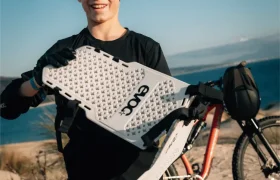 Un niño de pie sobre un terreno arenoso cerca de una playa, sosteniendo una tabla de tracción blanca con la etiqueta "EVOCO", con un EVOC Torso Protector talla XS para seguridad y una bicicleta de montaña al fondo.