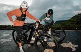Dos ciclistas con casco, talla XS EVOC Torso Protector y equipo de protección junto a sus bicicletas de montaña en una superficie pavimentada con dibujos de tortugas, al aire libre bajo un cielo nublado, dando prioridad a la seguridad durante su recorrido.