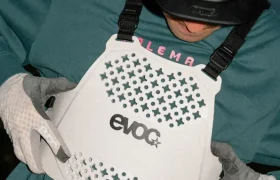 Una persona con casco y guantes ajusta un protector de torso EVOC blanco con diseño perforado, garantizando la seguridad mientras comprueba el ajuste de la talla XS.