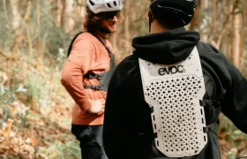 Dos ciclistas con casco y EVOC Torso Protector for seguridad de pie en una pista forestal, uno mirando a la cámara con una bicicleta, el otro girado hacia un lado hablando. Ambos parecen listos para la aventura, con equipo en talla XS.