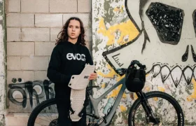 Una persona vestida de oscuro permanece de pie junto a una bicicleta, sosteniendo un EVOC Torso Protector talla XS para seguridad, frente a una pared desgastada con un gran mural de calaveras.