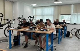 Personas sentadas en bancos de trabajo montando o reparando piezas de bicicleta en una sala luminosa con bicicletas, equipamiento ciclista y materiales de Formaciones Shimano 2026 a la vista.