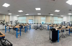 Amplio taller con mesas azules, bicicletas, piezas de ciclismo y herramientas organizadas por la sala bajo un entramado de luces de techo y grandes ventanales, ideal para Formaciones Shimano 2026.