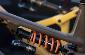 Primer plano de un sistema de suspensión trasera de bicicleta de montaña con un muelle helicoidal naranja y amortiguadores de alto rendimiento, con otra bicicleta con una configuración similar al fondo.