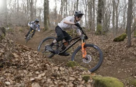Dos ciclistas de montaña con casco y amortiguadores FOX bajan por un sendero de tierra a través de un bosque con árboles desnudos y hojas caídas, mostrando la auténtica aventura del Factor X.