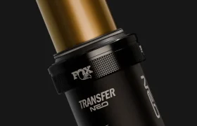 Primer plano de una tija telescópica electrónica FOX Transfer Neo E-Optimized para bicicleta de montaña, en el que se aprecia el tubo central dorado y la sección inferior negra con el logotipo sobre un fondo oscuro.