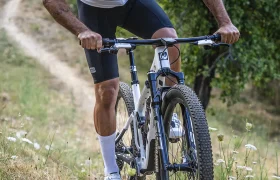 Un ciclista, que lleva casco y gafas de sol, sube en una bicicleta de montaña KFX i30 por un estrecho sendero de tierra que atraviesa una zona boscosa, manteniendo con destreza el control absoluto en este terreno tan exigente.