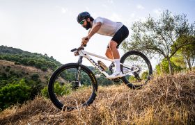 Un hombre, con casco y equipación de ciclismo, desciende en una bicicleta de montaña equipada con ruedas FSA por una colina seca y cubierta de hierba, manteniendo un control total mientras los árboles y las colinas se extienden al fondo.