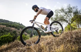 Una persona con casco baja en bicicleta de montaña, equipada con ruedas FSA KFX i30, por una colina seca y cubierta de hierba, demostrando un control total, con árboles y un cielo azul al fondo.