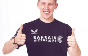 Un joven que viste una camiseta con la frase "Bahrain Victorious" sonríe y levanta los dos pulgares sobre un fondo blanco, encarnando el espíritu de equipo del Nutricionista Bahrain Victorious, como Giacomo Tomio.