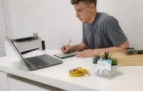 Un hombre está sentado en un escritorio blanco, escribiendo en un cuaderno mientras mira una laptop. Sobre el escritorio hay útiles de oficina, una pequeña planta y una caja; quizá tomando notas del nutricionista Bahrain Victorious, Giacomo Tomio.