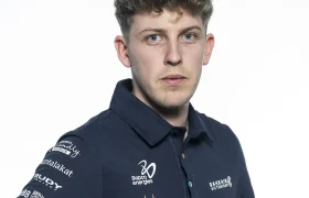 Un hombre joven con cabello corto y rizado que viste un polo azul marino con los logotipos de varios patrocinadores, incluido Nutricionista Bahrain Victorious, está parado sobre un fondo blanco liso.
