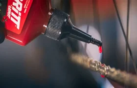 Una mano enguantada que aplica lubricante rojo para cadenas de bicicleta a una cadena desde un frasco dosificador.