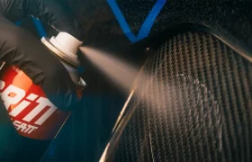 Una mano con un guante negro rociando un limpiador en aerosol sobre una superficie de fibra de carbono.