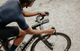 Un ciclista con casco y ropa azul conduce una bicicleta de carretera con un teléfono inteligente montado sobre una superficie de hormigón gris.