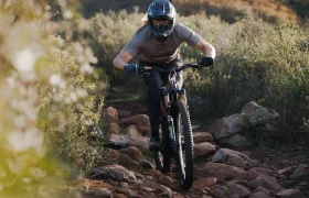 Persona con casco y guantes conduce una bicicleta de montaña Enduro con horquilla FOX 38 2027, manteniendo el control en un sendero rocoso rodeado de arbustos.