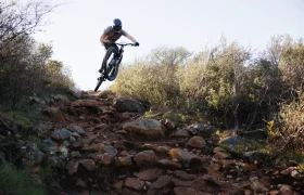 Un ciclista de montaña con casco y montado en una bicicleta de Enduro con horquilla FOX 38 2027 aborda un sendero rocoso, levantando la rueda delantera del suelo entre arbustos y árboles al fondo.