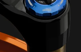 Primer plano de un mando de ajuste de horquilla de suspensión FOX 38 2027 con dial azul etiquetado "High Speed Compression" y "GRIPX2", que ofrece un control preciso de enduro en una horquilla de bicicleta negra y naranja.