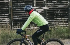 Una persona con casco y chaqueta brillante monta en bicicleta por un camino de tierra frente a un viejo edificio de madera desgastada, captando el espíritu de States of Elevation - Kilian Jornet.