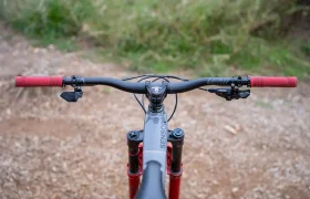 Vista aérea del manillar de una bicicleta de montaña, en la que destacan los puños ReaFlex 6.0 en un llamativo color rojo, las palancas de freno, y una palanca de cambio electrónica montada en un manillar Leatt Launch Control 6.0, con una potencia Gravity 6.0 que completa el conjunto; todo ello situado en un camino de tierra con una exuberante vegetación al fondo.