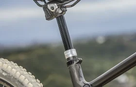 Primer plano de un sillín de bicicleta de montaña, una tija telescópica inalámbrica RAGE Circuit y una rueda trasera con un neumático cubierto de suciedad, sobre un fondo de paisaje borroso al aire libre.