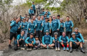Un grupo de ciclistas con maillots azules a juego posan juntos al aire libre, unos de pie y otros sentados, rodeados de árboles y vegetación seca, representando con orgullo a KS Sportmed y su distribución oficial.