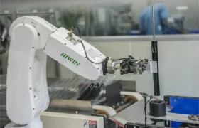 Un brazo robótico HIWIN sujeta un objeto cilíndrico en un entorno de fabricación o laboratorio, con KS como distribución oficial en España; al fondo se ve maquinaria y una persona.