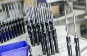 Filas de componentes metálicos y negros parcialmente ensamblados, posiblemente amortiguadores o cilindros neumáticos, se alinean en un entorno de fábrica o taller con contenedores azules cerca, destacando la distribución oficial KS España de Sportmed.