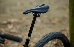 Primer plano de un sillín y una tija de bicicleta KS negros con un neumático trasero de tacos, tomados al aire libre cerca de un árbol con un fondo borroso y terroso. Distribución oficial Sportmed.