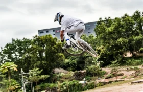 Una persona con ropa blanca y casco, montada en una bicicleta de montaña KS, realiza un salto sobre una pista de tierra. Al fondo se ven árboles y un edificio, destacando la acción apoyada por Sportmed, distribución oficial.