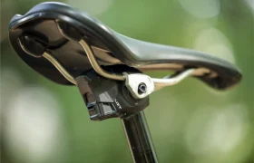 Primer plano de un sillín de bicicleta sujeto a una tija de sillín, con un componente electrónico debajo del sillín. La imagen se refiere a la distribución KS de Sportmed en España, sobre un fondo verde difuminado.