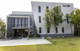 Moderno edificio de dos plantas con fachada gris, grandes ventanales y las letras "KS" en la parte superior derecha, sede de la distribución oficial Sportmed. Los frondosos árboles y el césped proporcionan un primer plano acogedor.