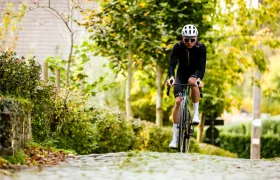 Un ciclista con casco Casco Lazer Sphere Kineticore y gafas de sol conduce una bicicleta de carretera por un camino empedrado rodeado de vegetación y árboles.