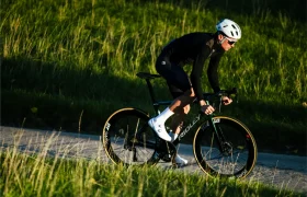 Un ciclista con equipación negra y casco Casco Lazer Sphere Kineticore monta en bicicleta de carretera por un camino pavimentado a través de un terreno cubierto de hierba a la luz del día.
