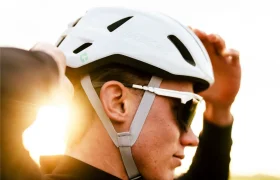 Una persona con gafas de sol ajusta un casco Casco Lazer Sphere Kineticore blanco, con la luz del sol brillando en el fondo.