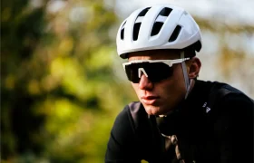 Una persona con un casco Casco Lazer Sphere Kineticore blanco, gafas de sol negras y un maillot negro de ciclista está al aire libre con vegetación borrosa de fondo.