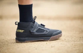 Una persona que lleva zapatillas negras Leatt de ciclismo con una tira de velcro se para sobre una superficie arenosa.