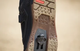 Primer plano de la suela de una zapatilla de ciclismo Leatt negra con una cala metálica visible, llevada por una persona que camina sobre una superficie arenosa.
