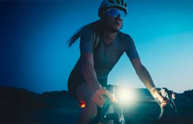Un ciclista con casco y gafas de sol monta en bicicleta con una luz frontal Lezyne Radar React System encendida al anochecer o al amanecer, con un paisaje oscuro de fondo.