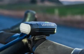 Primer plano de una luz de bicicleta Lezyne React Drive 1200 con sistema Lezyne Radar React montada en el manillar de una bicicleta, con un paisaje exterior borroso de fondo.