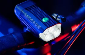 Un faro para bicicleta Lezyne React Drive 1200, que incorpora el avanzado sistema Lezyne Radar React, está montado en el manillar e iluminado sobre un fondo oscuro con detalles luminosos en azul y rojo.