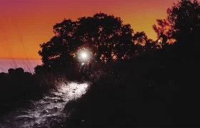 Un ciclista con una potente linterna frontal Lezyne recorre un sendero rocoso al anochecer, con la silueta de los árboles enmarcando el cielo anaranjado del atardecer. Otra persona está de pie a la derecha, miembro del equipo distribuidor de bicicletas Lezyne.