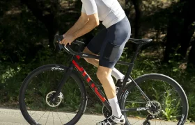 Un ciclista que lleva un casco blanco y un maillot de POC Cadence circula en una bicicleta de carretera negra y roja por una carretera asfaltada, disfrutando de la comodidad al pedalear, con árboles y vegetación al fondo.