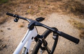 Manillar blanco de bicicleta de montaña, concretamente un manillar integrado de carbono FSA NS SIC, con un ciclocomputador montado en el centro, fotografiado al aire libre en un camino de tierra.