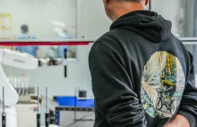 Un hombre con una sudadera negra con un gráfico de ciclista está de pie con las manos a la espalda, observando el equipo de tija telescópica inalámbrica RAGE Circuit en un laboratorio o taller moderno.