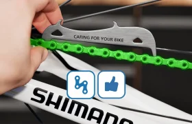 Una mano utiliza un medidor de cadena en una cadena de bicicleta verde, con la palabra "SHIMANO" visible en el cuadro, asegurándose de que la cadena original Shimano se comprueba para ver si está desgastada.