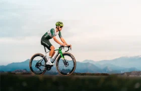 Un ciclista vestido de verde y blanco conduce una bicicleta de carretera MMR Aelion SL por un sendero con montañas y un cielo nublado de fondo.