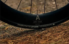 Primer plano de una rueda de bicicleta que muestra el vástago de la válvula, la llanta negra con la etiqueta "ARC 1600" y el neumático con la marca "Hutchinson Blackbird", con agujas de pino y gravilla en el suelo: perfecta para tu próximo paseo con la MMR Aelion SL de bici total.